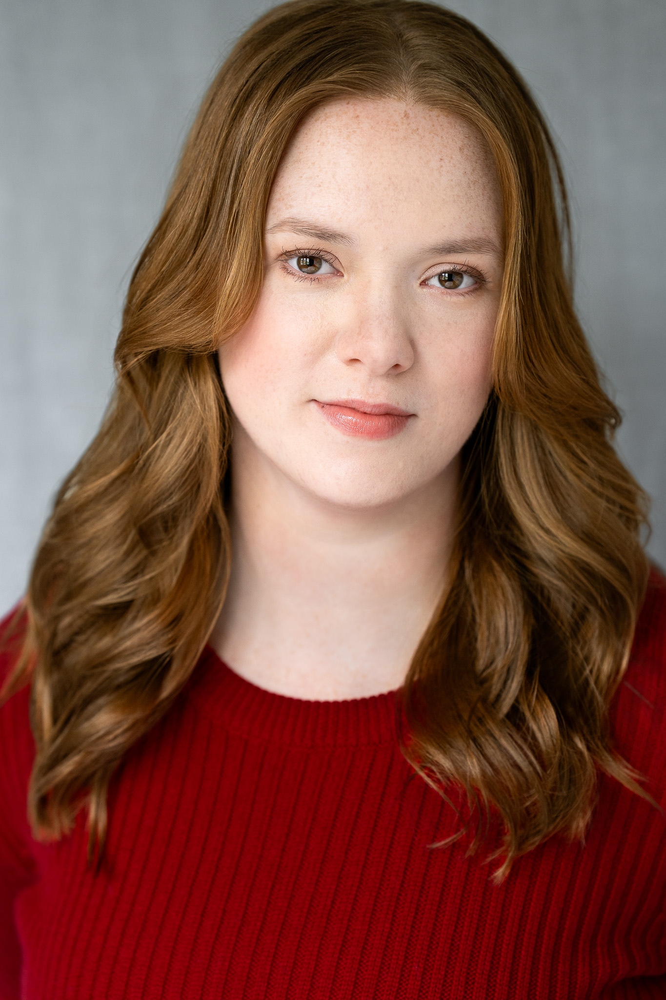 Young woman with long, curled auburn hair, light freckles, wearing a red knit sweater, smiling softly against a gray background.