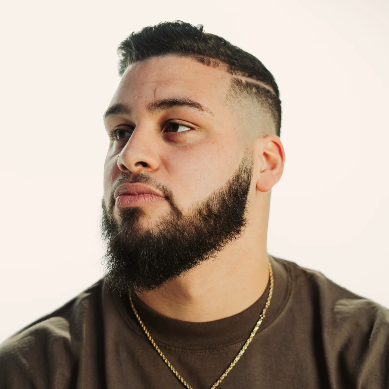 Bearded man with a short haircut and a line shaved on the side, wearing a brown shirt and gold chain, looking to the left.