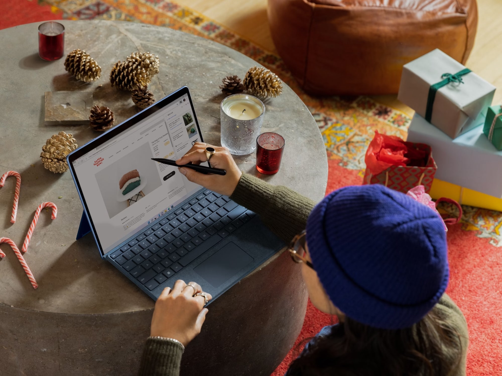 Person in blue knit hat shopping online for sweaters on a laptop at a round table with pinecones, candles, candy canes, and holiday gifts nearby.