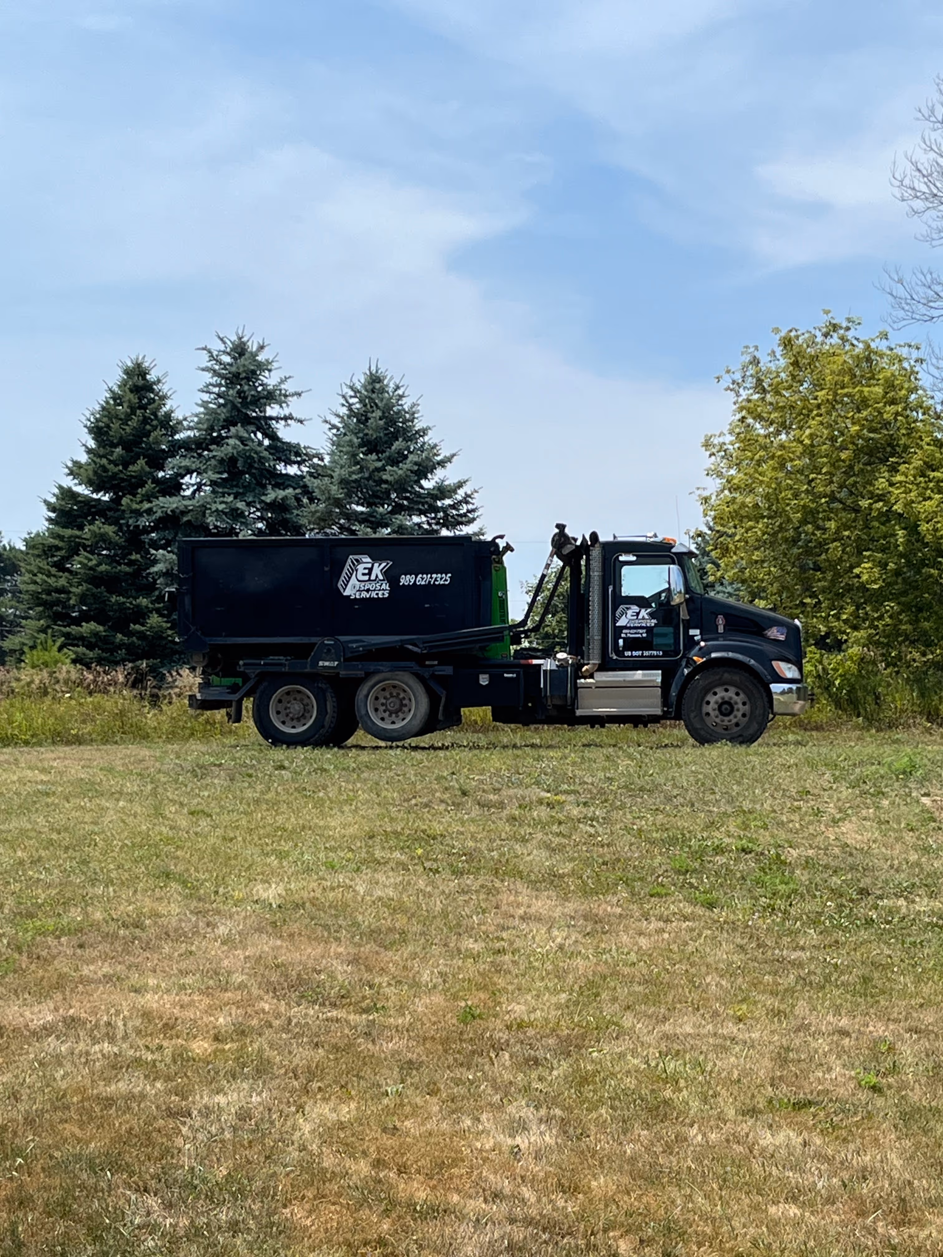 Black EK Disposal Services roll-off dumpster truck parked on grassy field with trees in the background.