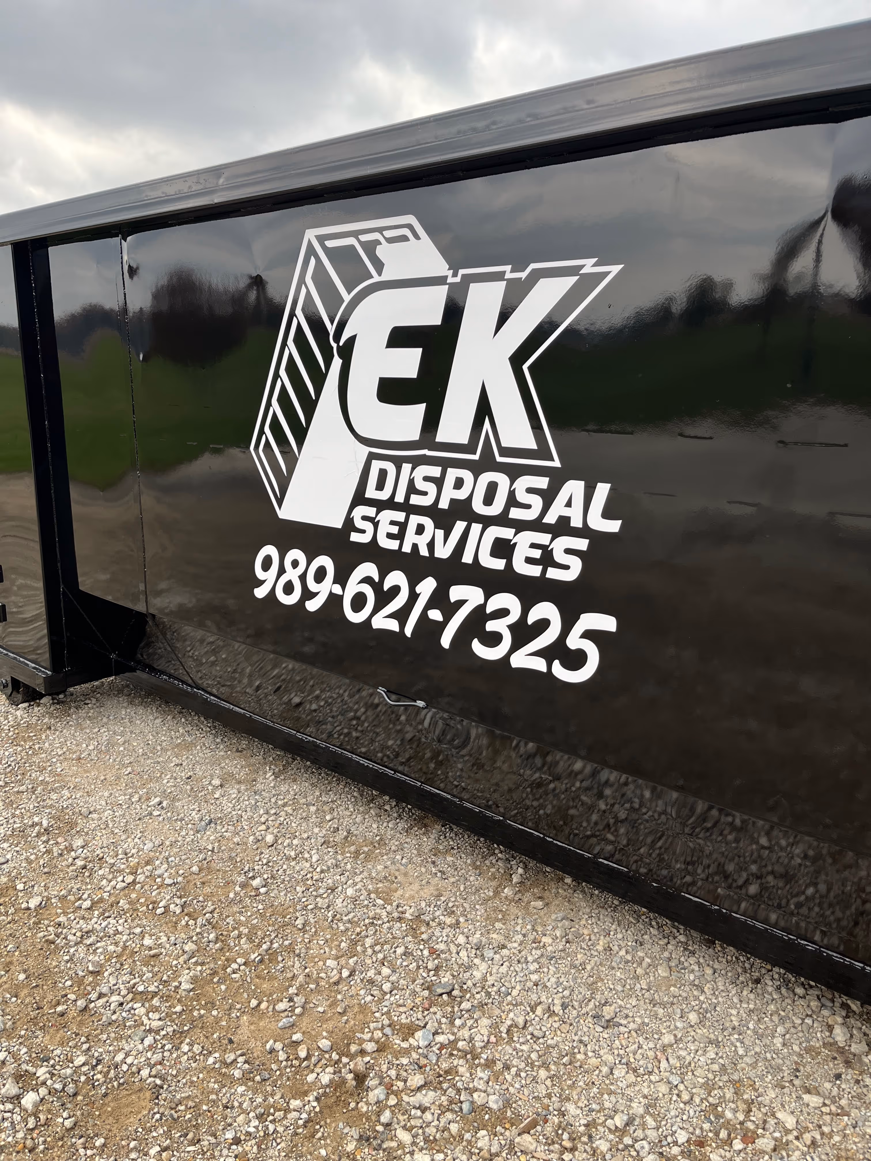 Black disposal dumpster with white EK Disposal Services logo and phone number 989-621-7325 on its side.