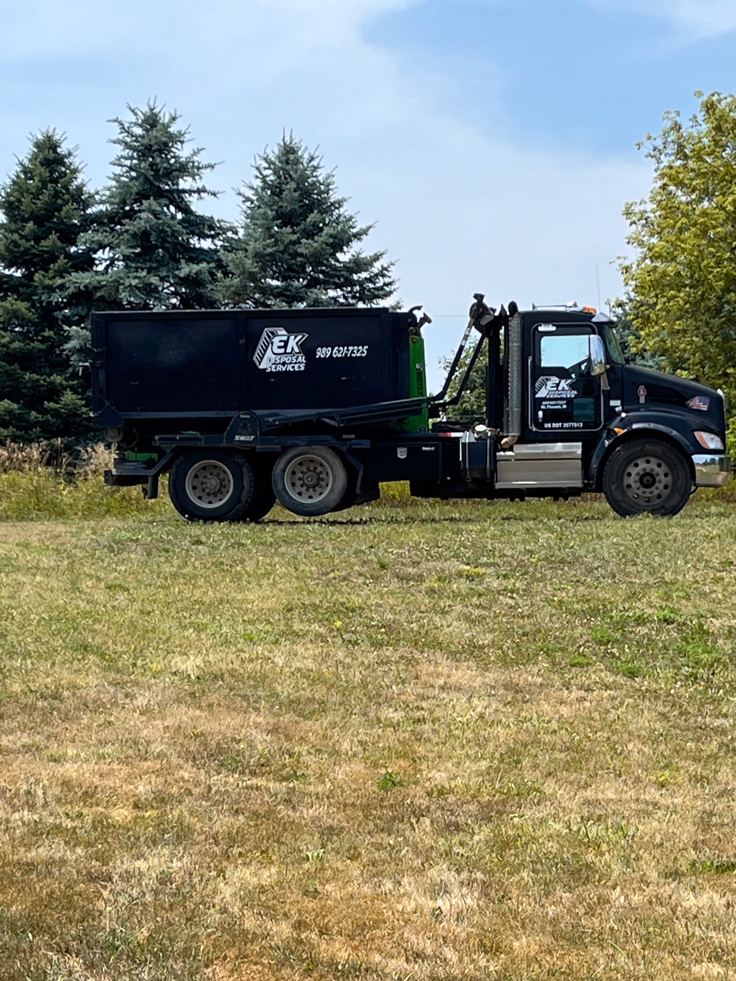 Black EK Disposal Services roll-off truck parked on a grassy field with evergreen trees in the background.