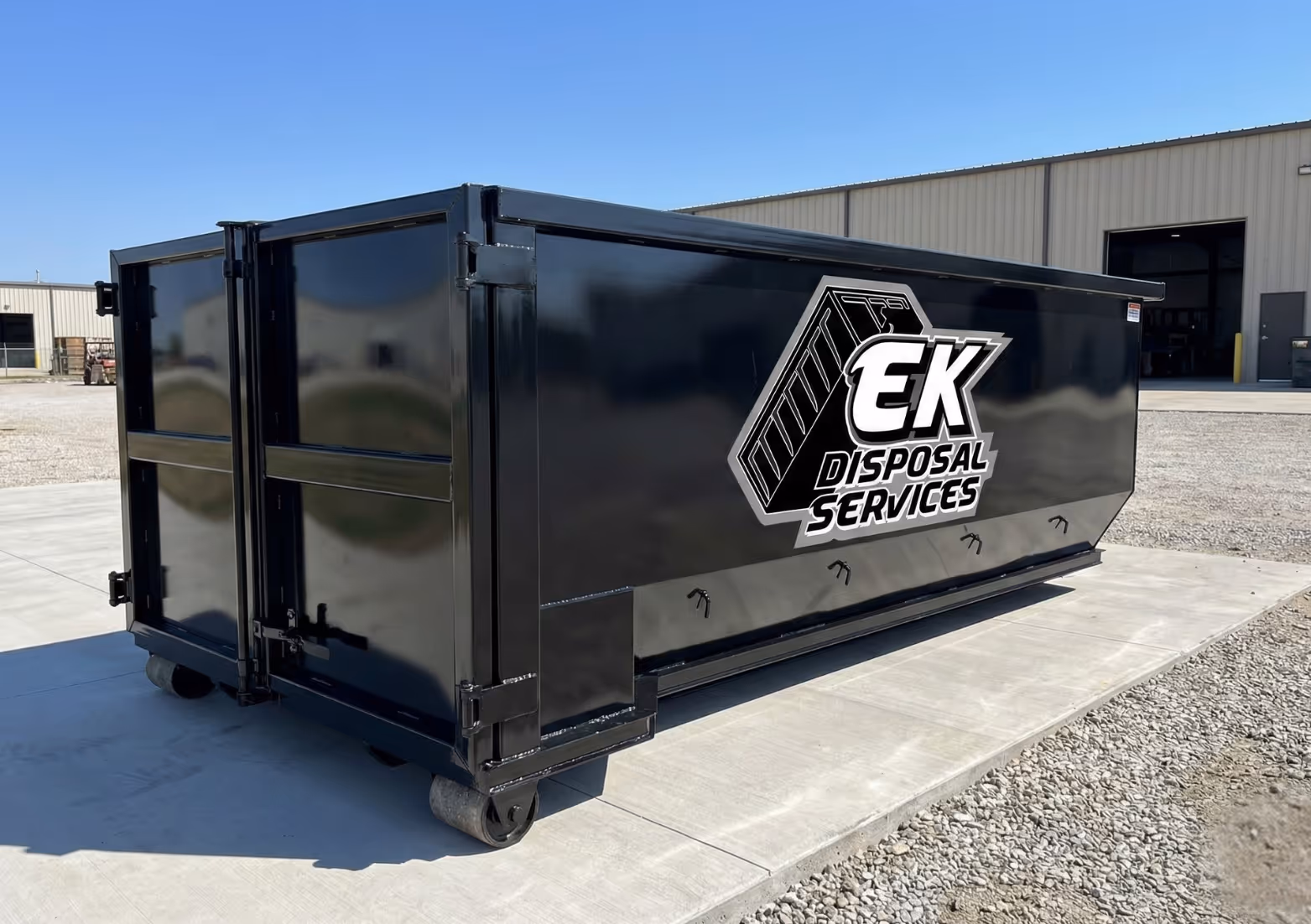Large black industrial dumpster with EK Disposal Services logo on the side, placed on a concrete pad outside a warehouse.