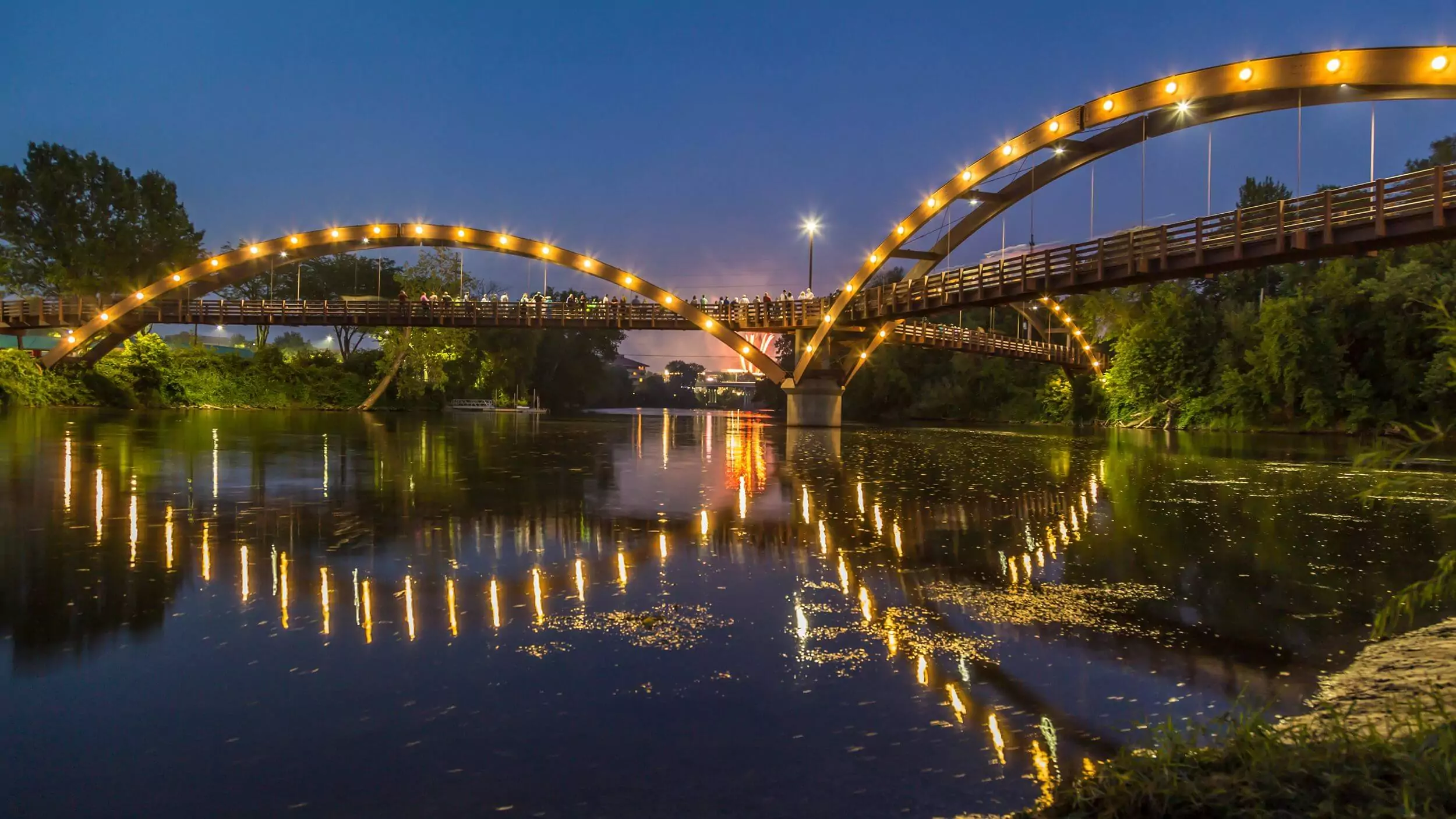 Illuminated pedestrian bridge with arches reflecting on calm river water during twilight.