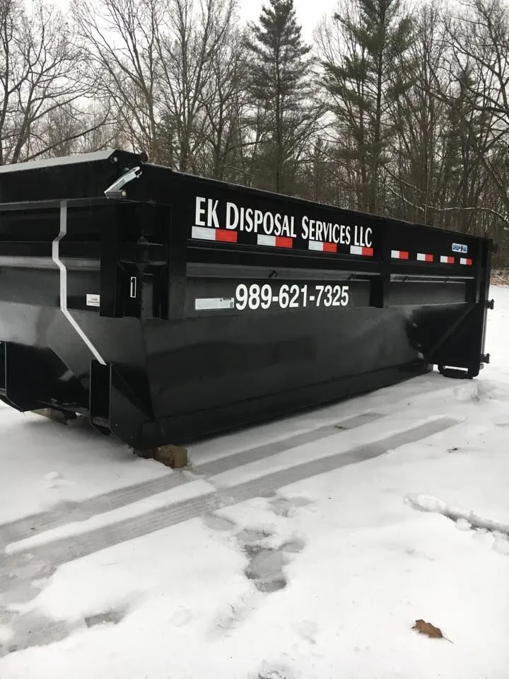 Black disposal container with 'EK Disposal Services LLC' and phone number 989-621-7325 printed on the side, placed on snow-covered ground with leafless trees in the background.