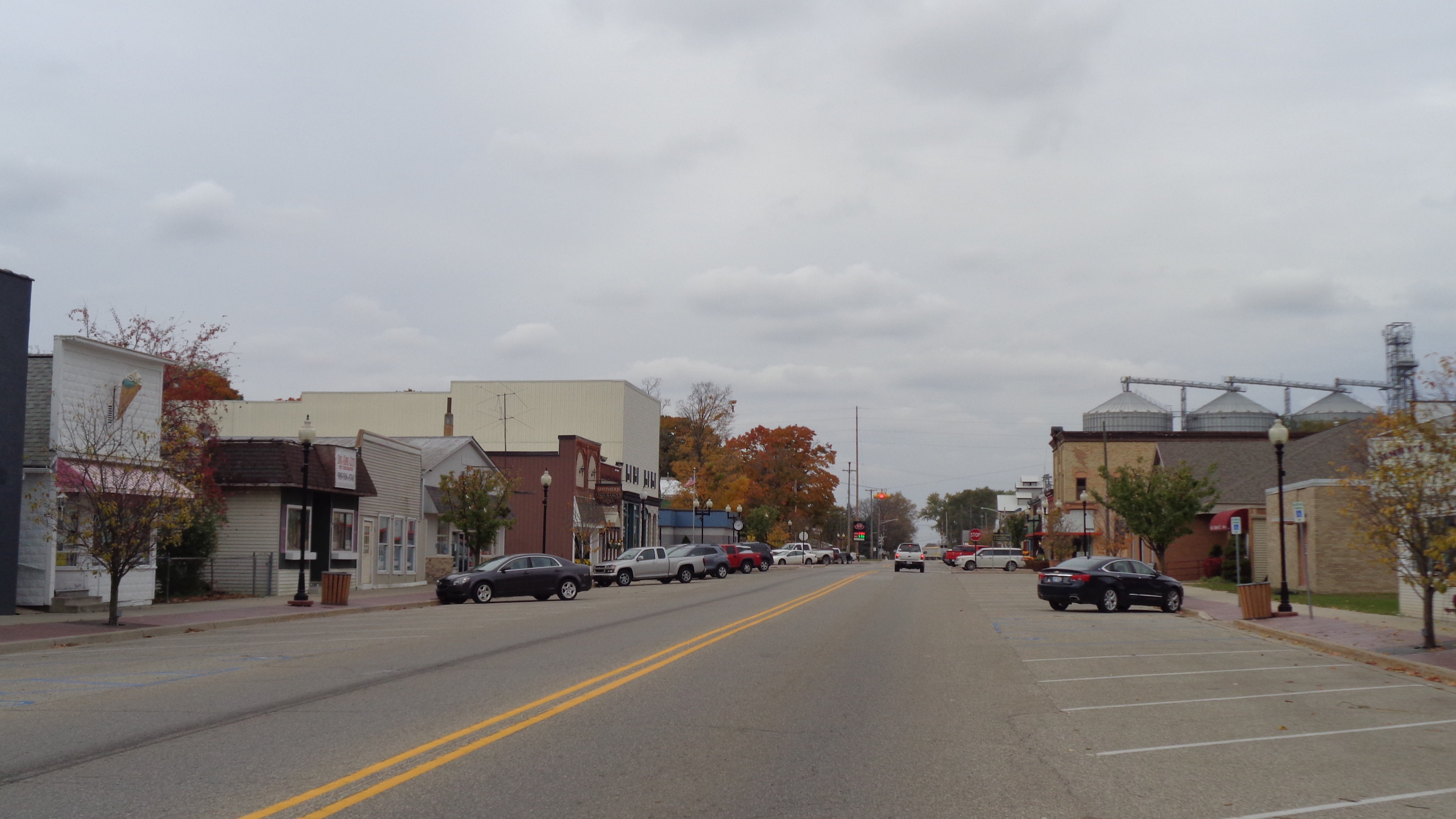 Empty main street in a small town with parked cars and buildings under a cloudy sky.
