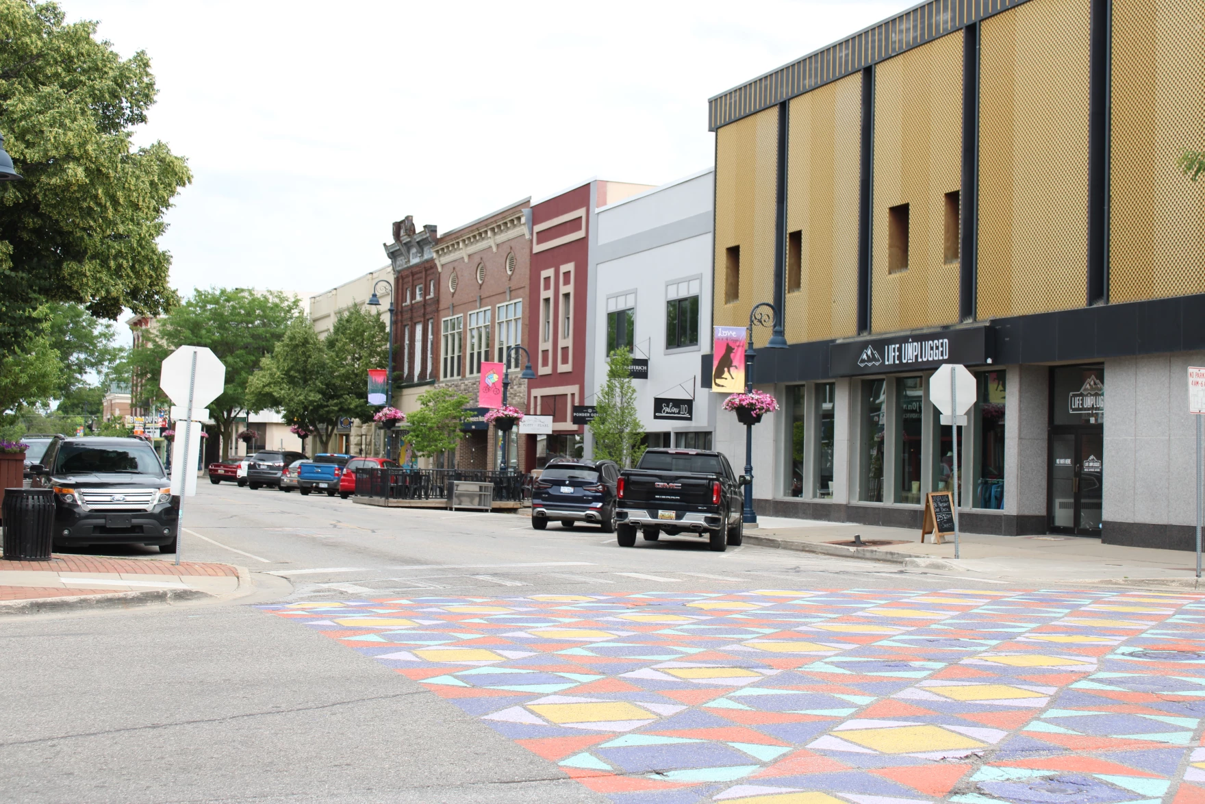 Downtown street with parked cars, colorful geometric crosswalk, and a row of storefronts including Life Unplugged.