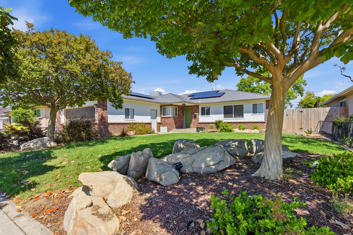 Single-story house with solar panels on roof, brick and white exterior, lush front yard with trees and rocks.