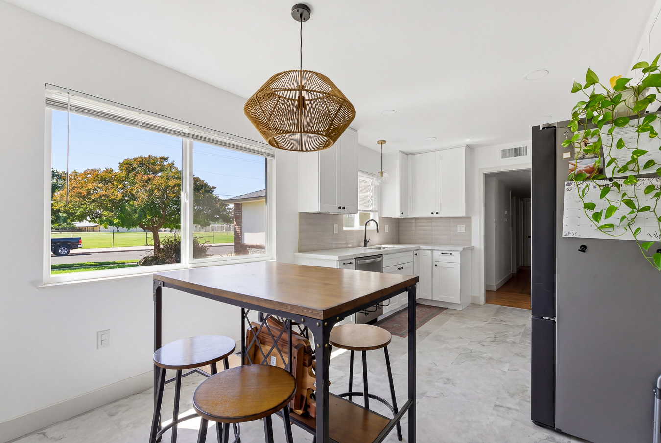 Bright kitchen with white cabinets, a wooden island table with stools, a rattan pendant light, and a large window.