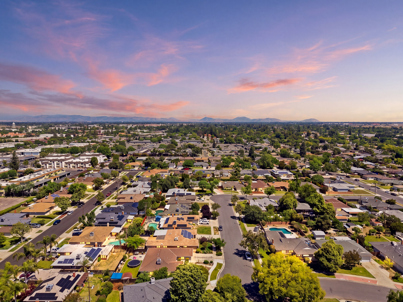 Aerial view of a suburban neighborhood with houses, trees, swimming pools, and a sunset sky.
