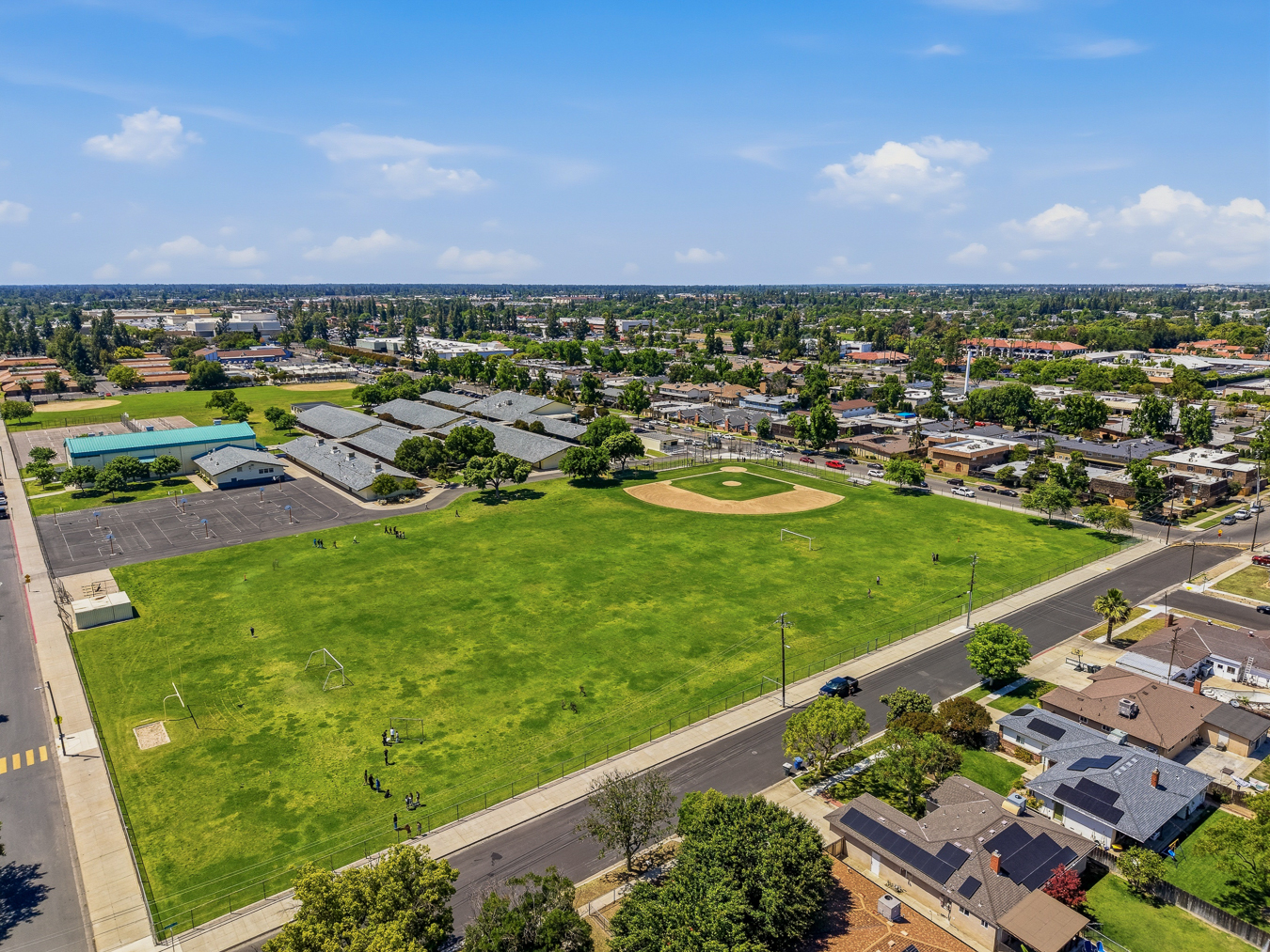 Aerial view of a school with basketball courts, a baseball field, green sports fields, and surrounding neighborhood.