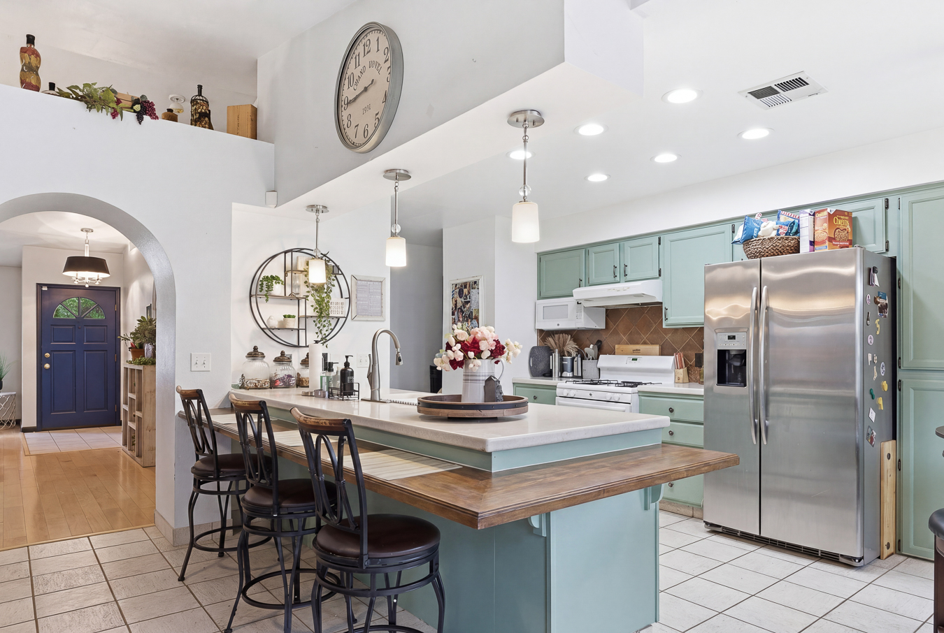 Kitchen with green cabinets, stainless steel fridge, island with chairs, pendant lights, and blue front door in background.