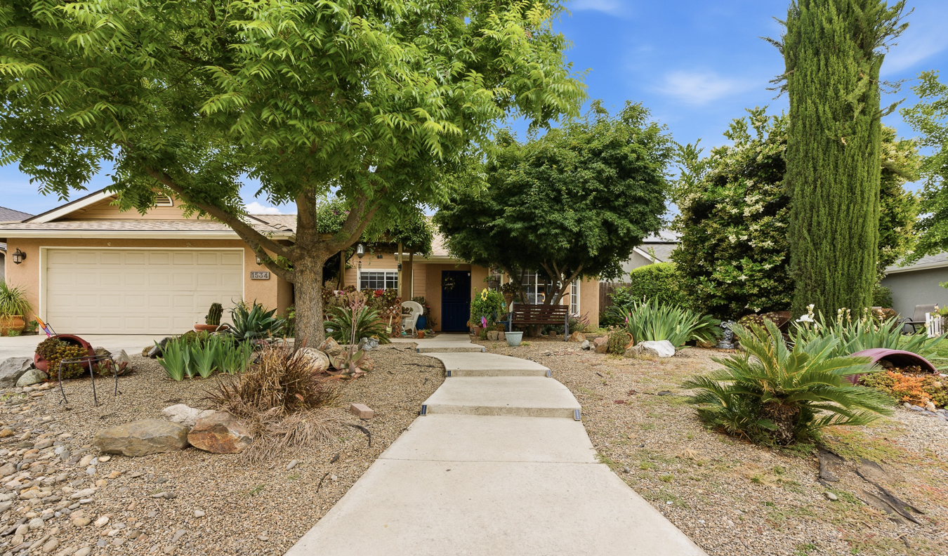 Front yard with concrete walkway, trees, shrubs, and a beige house with a two-car garage.