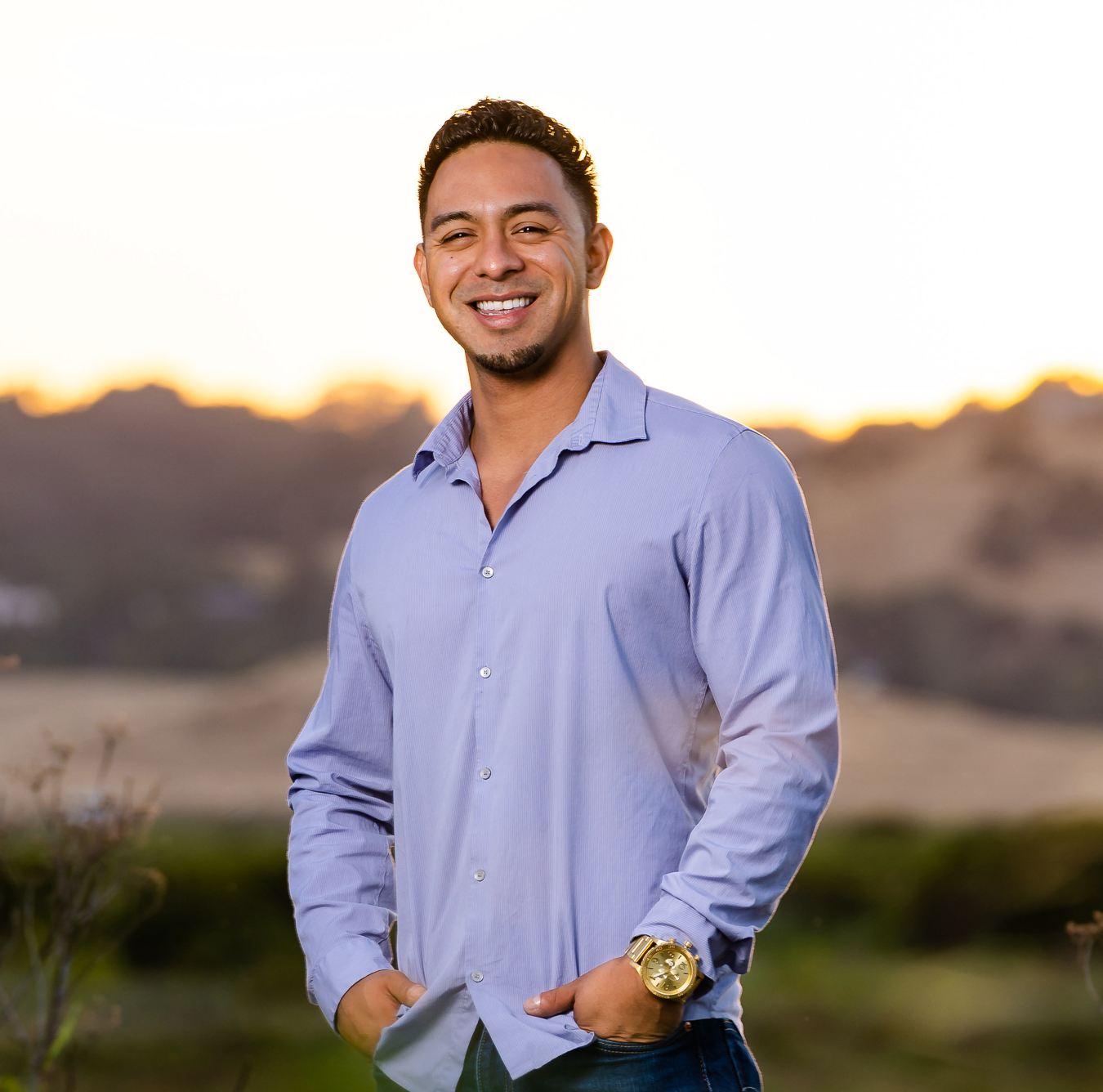 Smiling man in blue button-up shirt standing outdoors at sunset with hands in pockets.