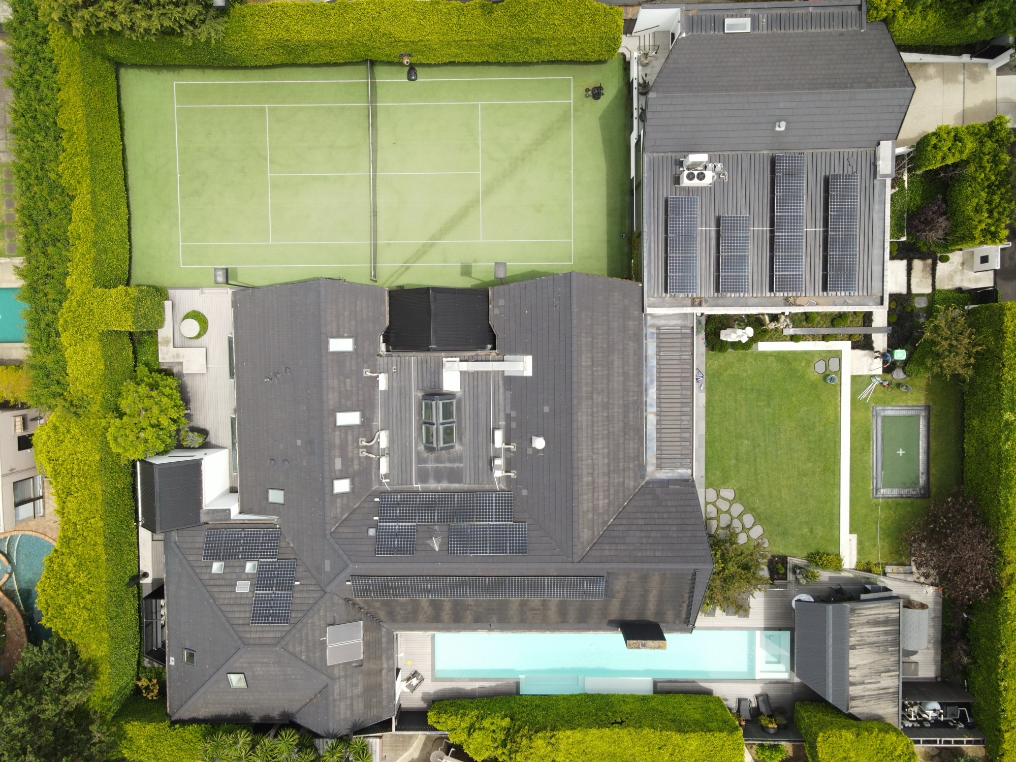 Aerial view of a modern house with solar panels, a tennis court, a rectangular swimming pool, and a manicured garden enclosed by tall green hedges.