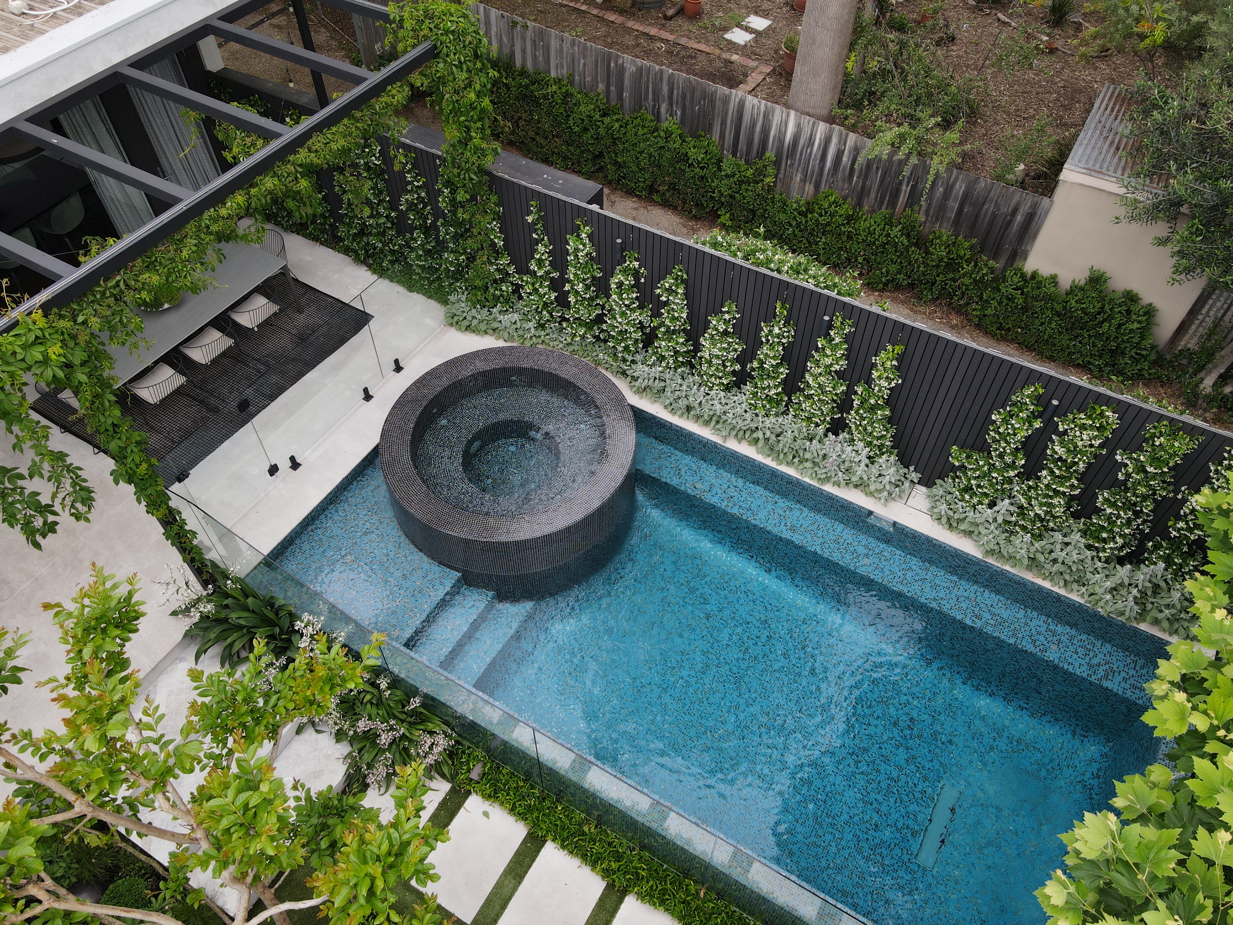 Modern outdoor pool with an integrated circular hot tub, surrounded by glass fencing and greenery.