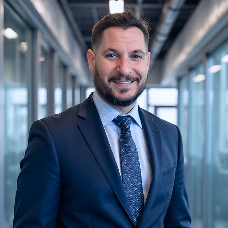 Smiling man with short dark hair and beard wearing a dark suit and blue tie in a modern office hallway.