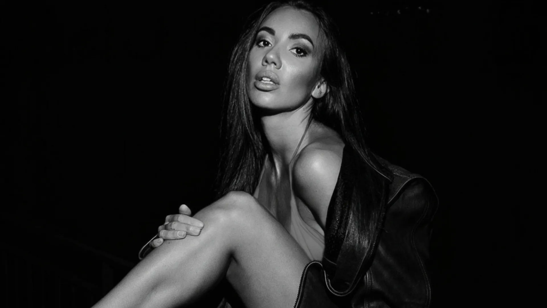 Black and white photo of a woman with long hair sitting, holding her leg, looking at the camera.