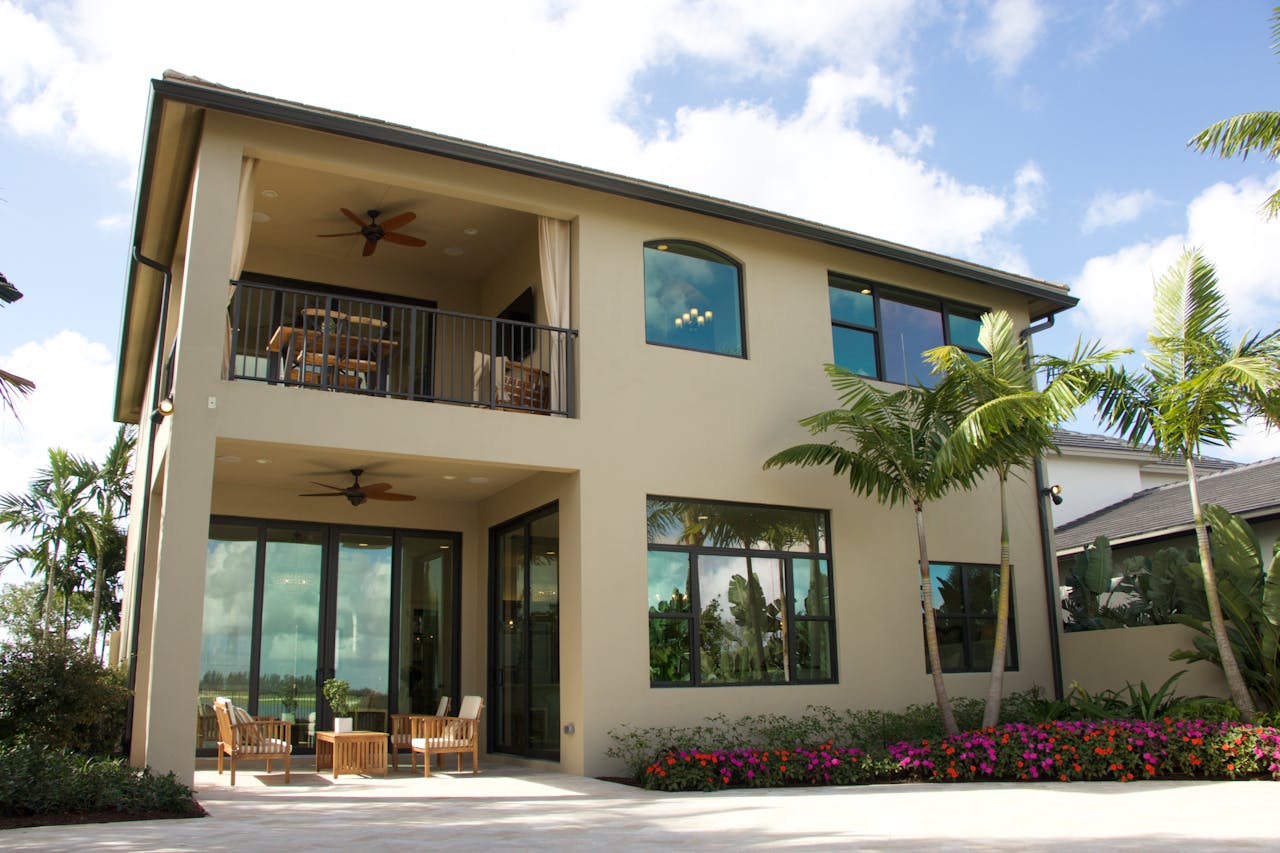 Modern two-story Florida home with flat dark roof, large glass windows, upper balcony with ceiling fan, covered patio with outdoor furniture, and tropical landscaping with colorful flowers