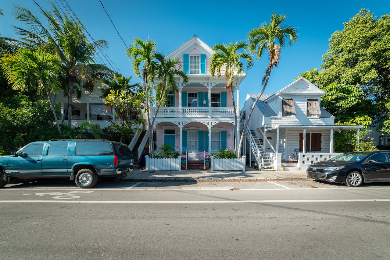 Colorful three-story Victorian-style home with teal shutters and wrap-around balcony on a Florida street lined with palm trees, parked vehicles visible in the foreground