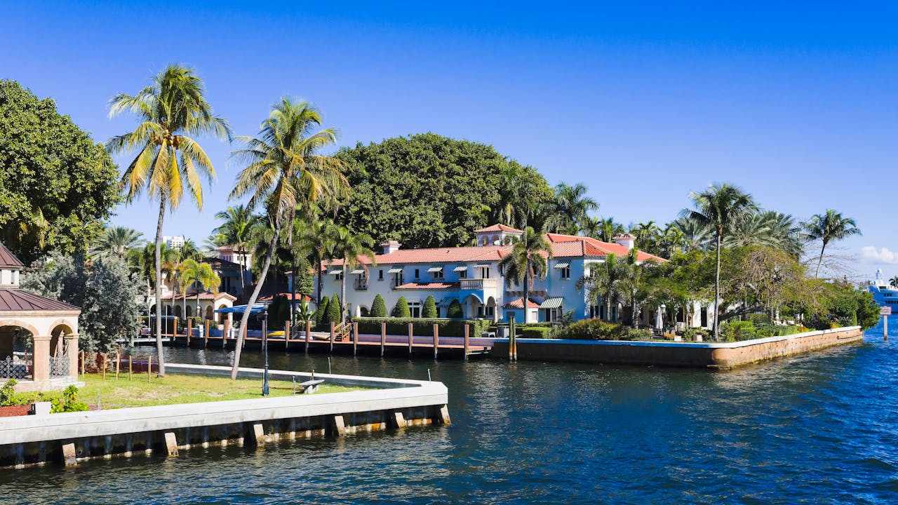 Waterfront Florida estate with terracotta tile roof, white stucco walls, and palm trees lining a calm canal on a clear sunny day