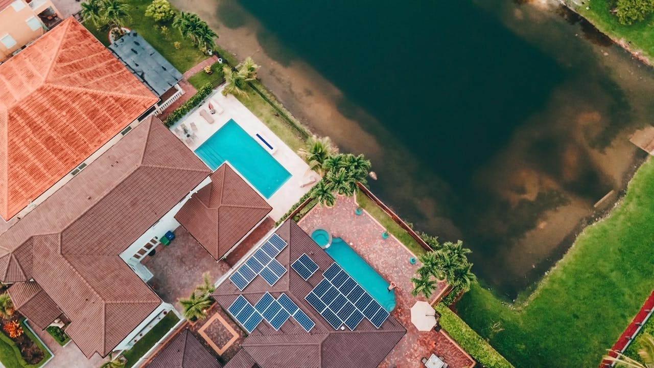Aerial drone view of a Florida residential property with terracotta tile roofs, solar panels installed on one section, a backyard swimming pool, and a lake visible in the background