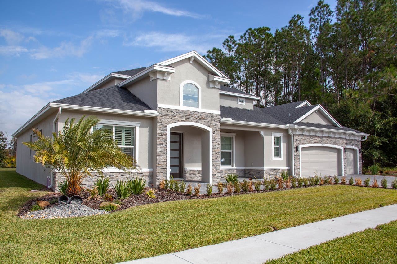 Single-story Florida home with gray shingle roof, stone veneer facade, attached two-car garage, palm trees, and manicured front lawn under a partly cloudy sky