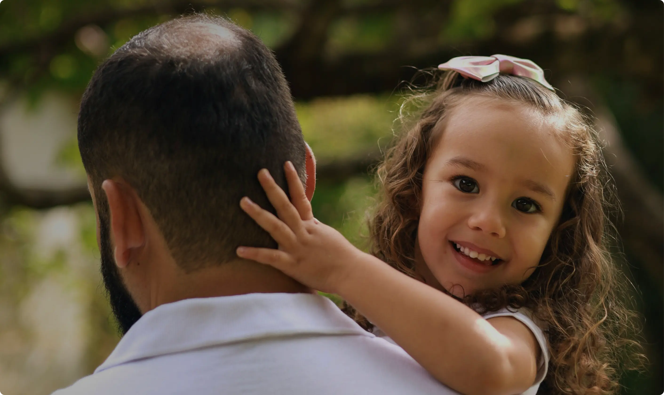 A young child smiling while being held over an adult’s shoulder outdoors.