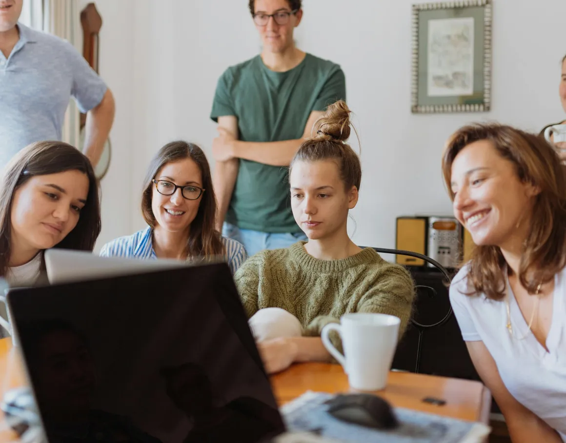 A group of people gathered around a laptop, collaborating and smiling at a table.