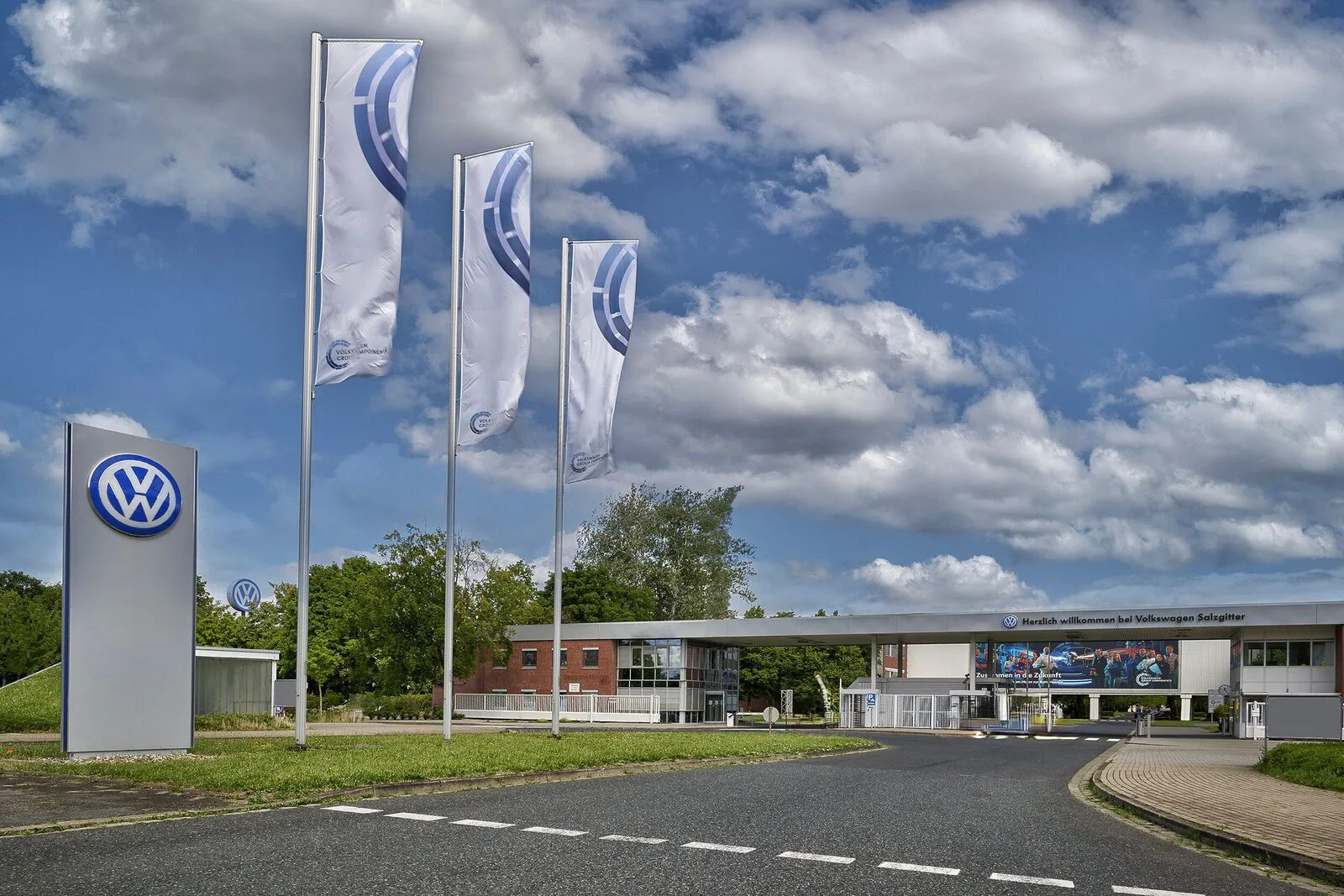 Vhod v obrat Volkswagen Salzgitter z značko Volkswagen, tremi zastavami in modro nebo z oblaki.