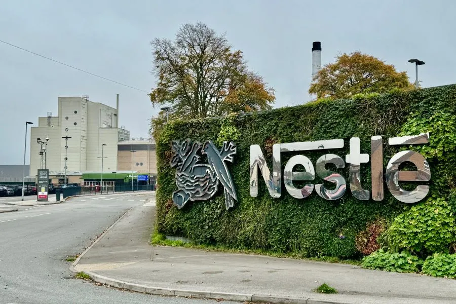 Napis Nestlé z zrcalnim učinkom na zelenem živem zidu ob cesti pred industrijskim objektom.