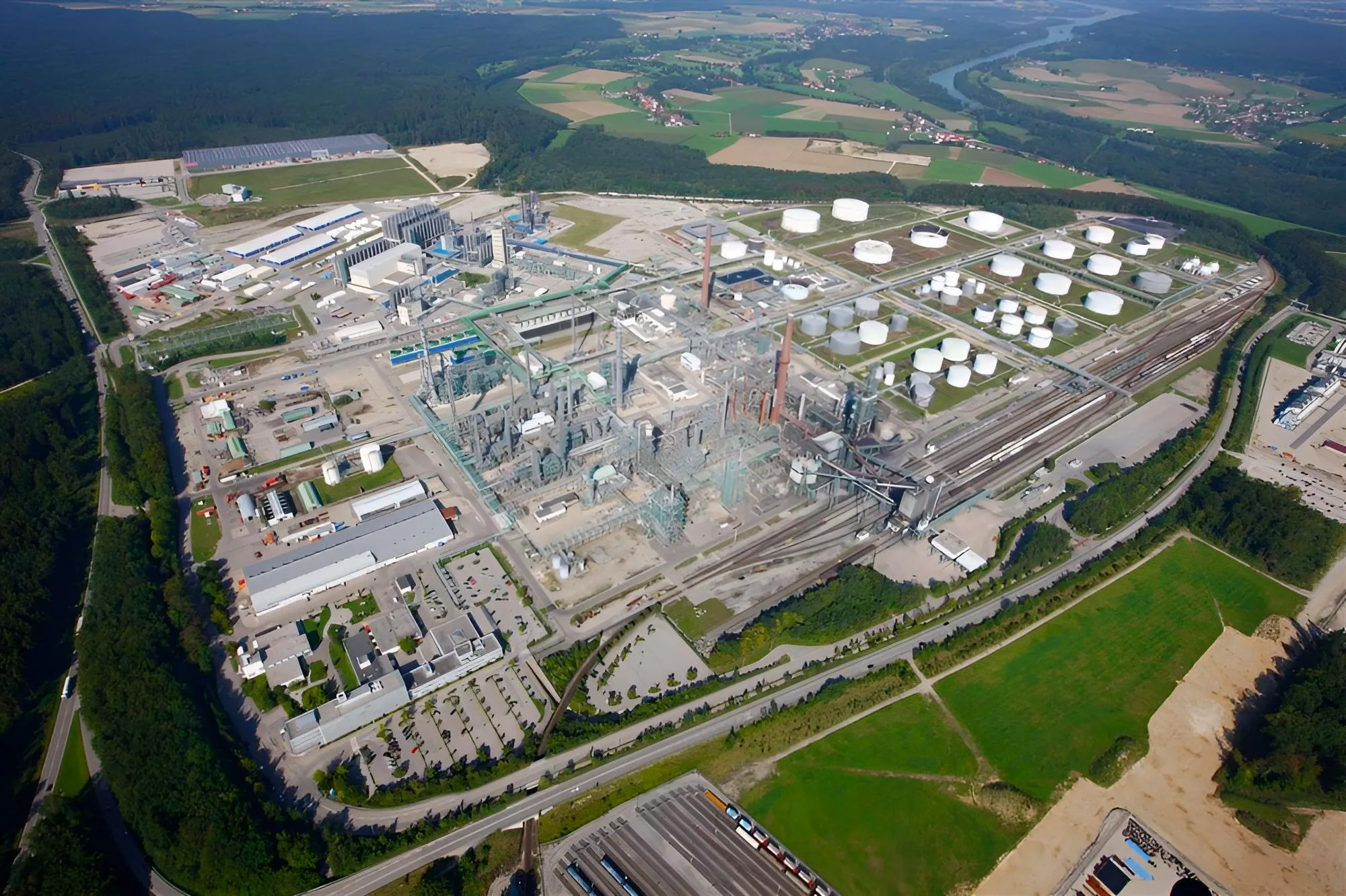 Pogled iz zraka na veliko industrijsko obrat z mnogimi skladiščnimi rezervoarji in železniško infrastrukturo, obdan z gozdom in kmetijskimi zemljišči.