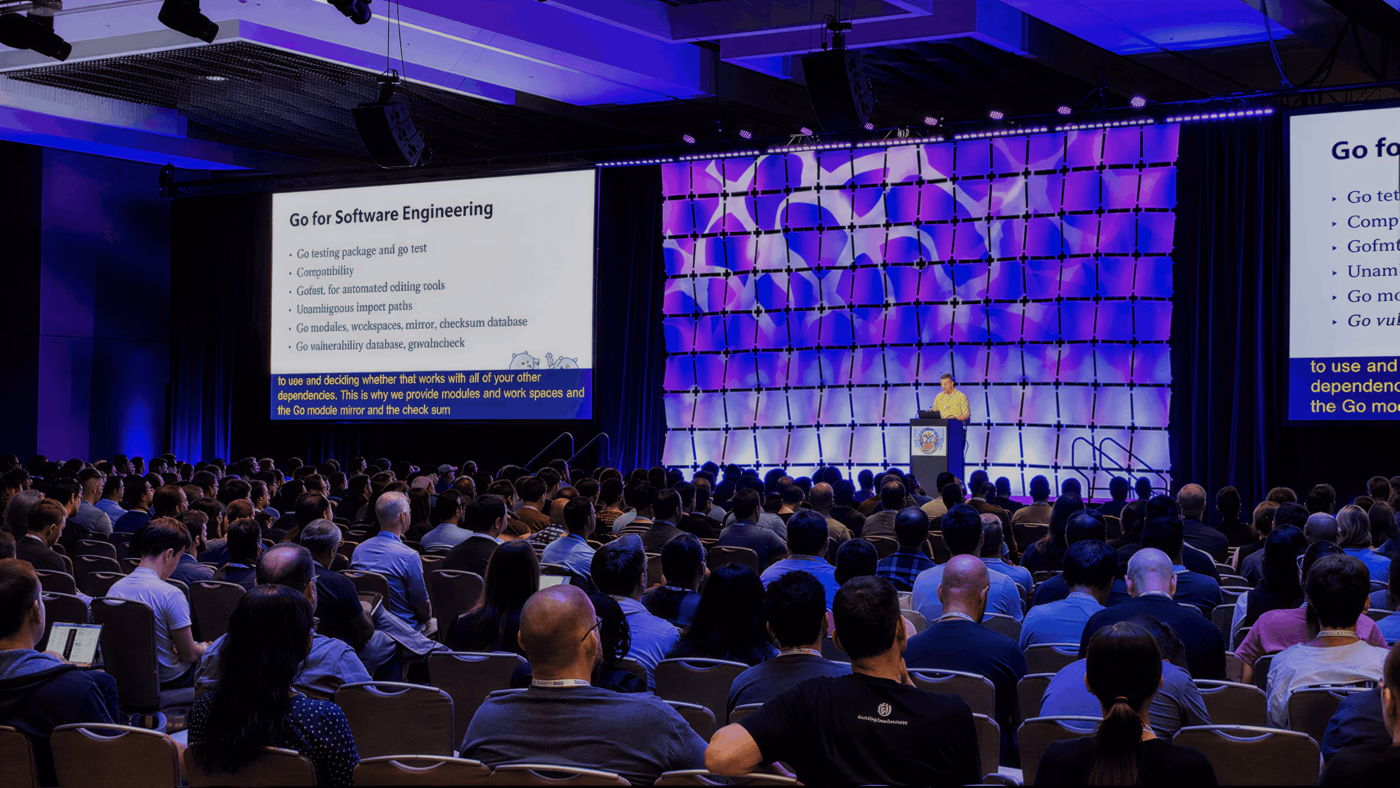 Audience seated in a conference room attending a presentation on software engineering with purple lighting and slides displayed on two large screens.