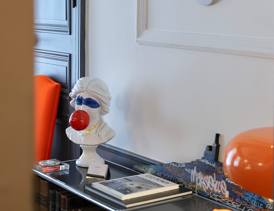 White bust sculpture with painted blue eyeshadow and yellow necklace blowing a large red bubble, placed on a glass table with artistic objects and books.