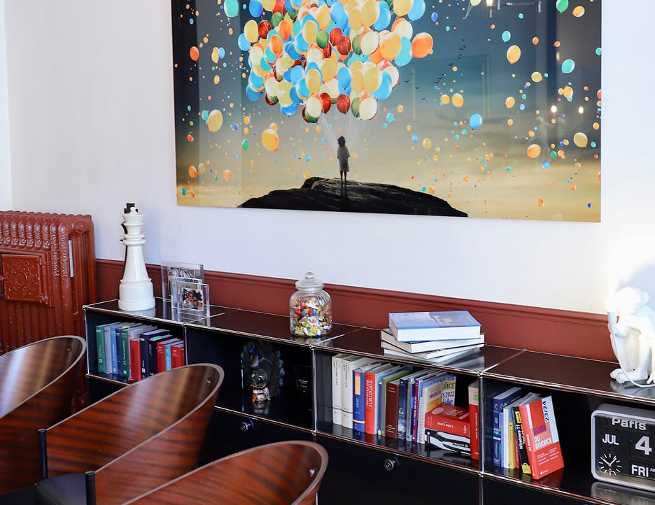 Modern office corner with wooden chairs, a black cabinet filled with books, a jar of colorful paper clips, a decorative white chess piece, and a large wall art showing a silhouette of a girl holding many colorful balloons.
