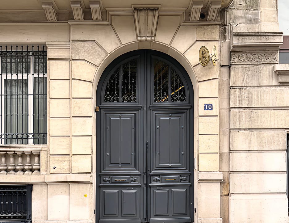 Large ornate dark gray double door with decorative glass panels above set in a beige stone building facade showing the number 10 beside the entrance.