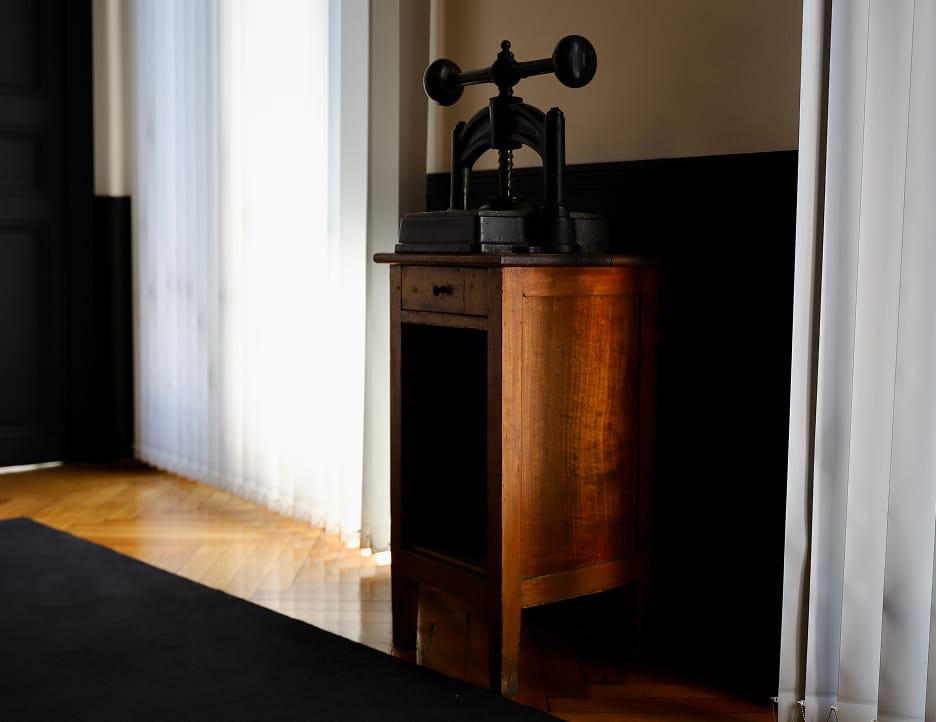 Antique wooden side table with a black vintage book press on top, next to white curtains and a window.