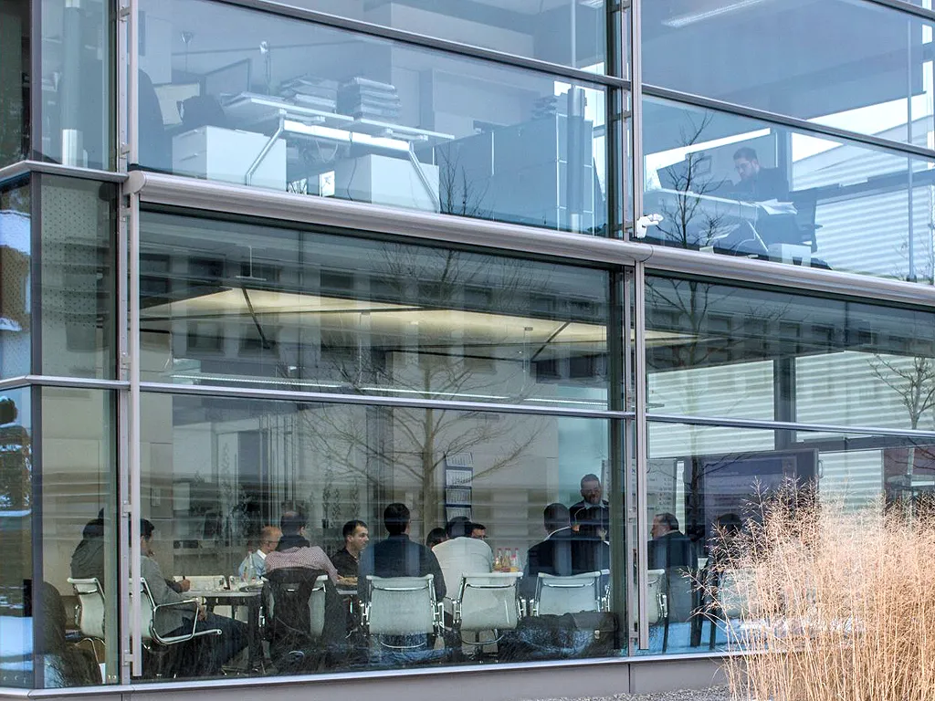 People seated around a conference table in a modern glass-walled office building, viewed from outside through large windows.