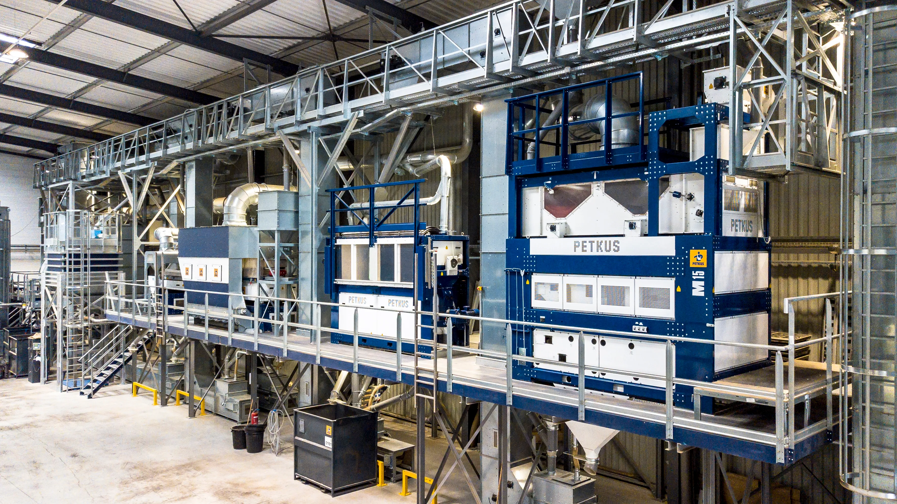 Large industrial machine setup inside a spacious warehouse with metal framework, ductwork, and multiple units labeled PETKUS, elevated on a platform with railings.