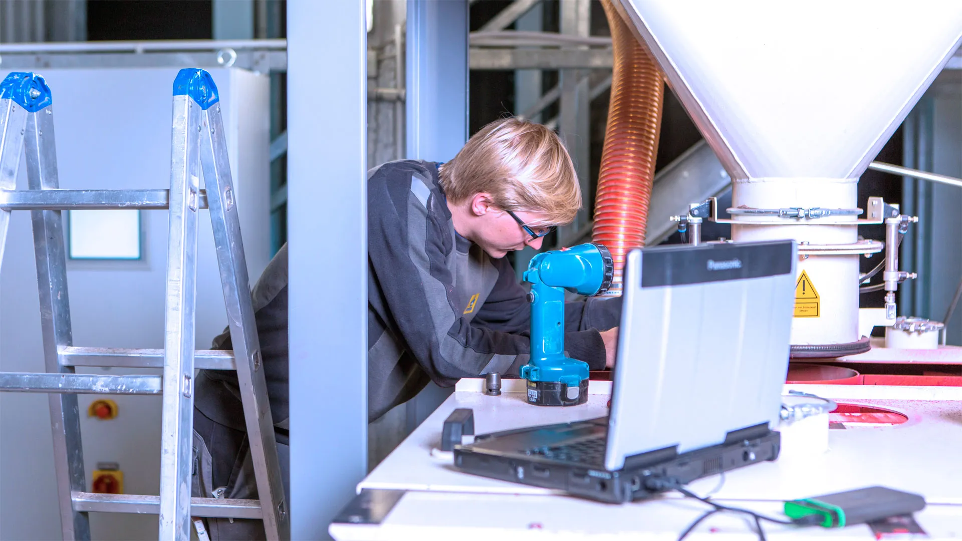 A technician with glasses and blond hair is working on industrial equipment indoors, leaning forward behind a workbench with a blue cordless drill and an open Panasonic laptop nearby, with a metal ladder to the left and a large white funnel-shaped component in the background.