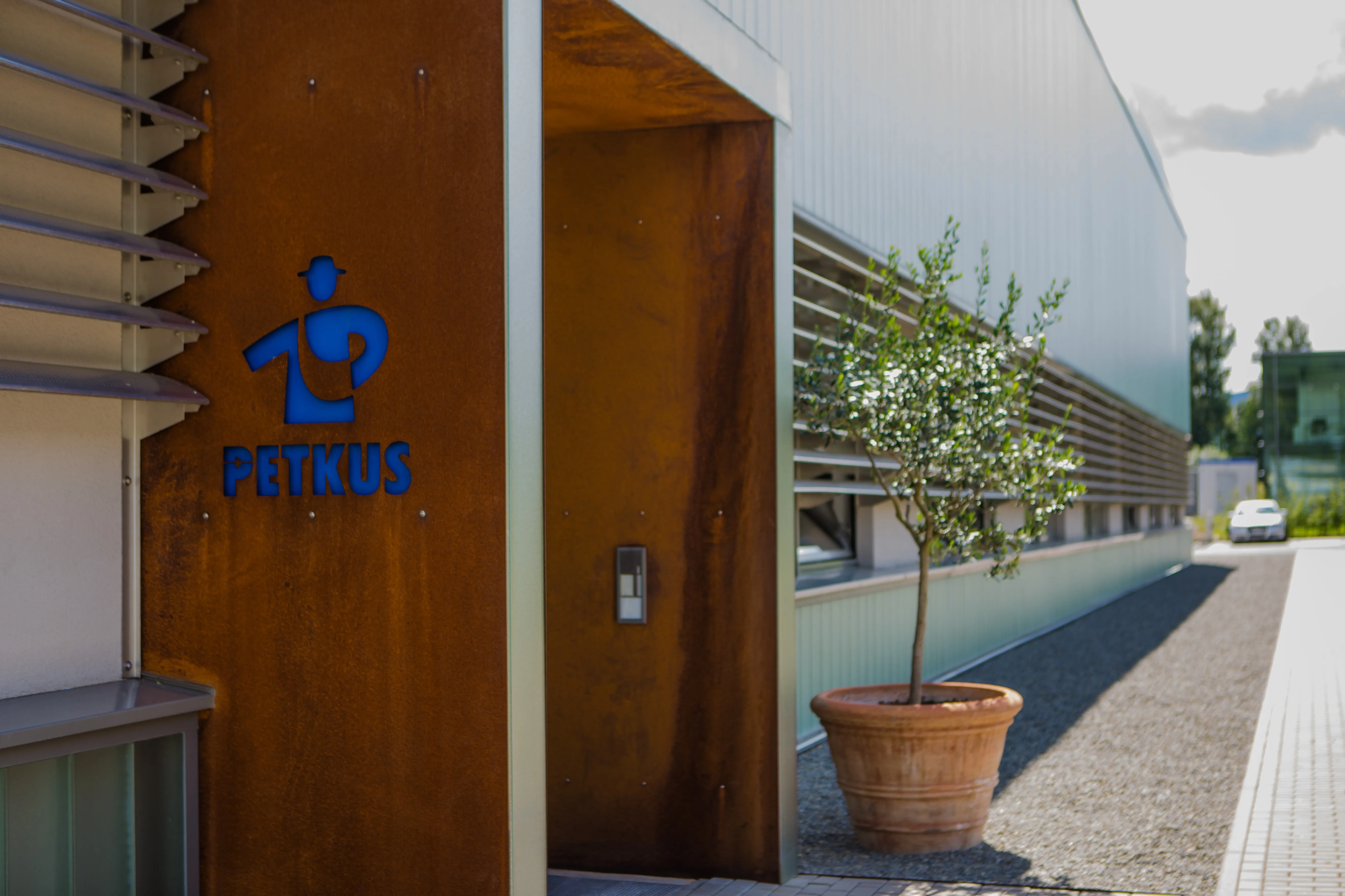 Entrance of a modern industrial building with a rust-colored metal panel featuring a blue PETKUS logo to the left, a potted tree on the right, and the building's exterior wall extending into the background.