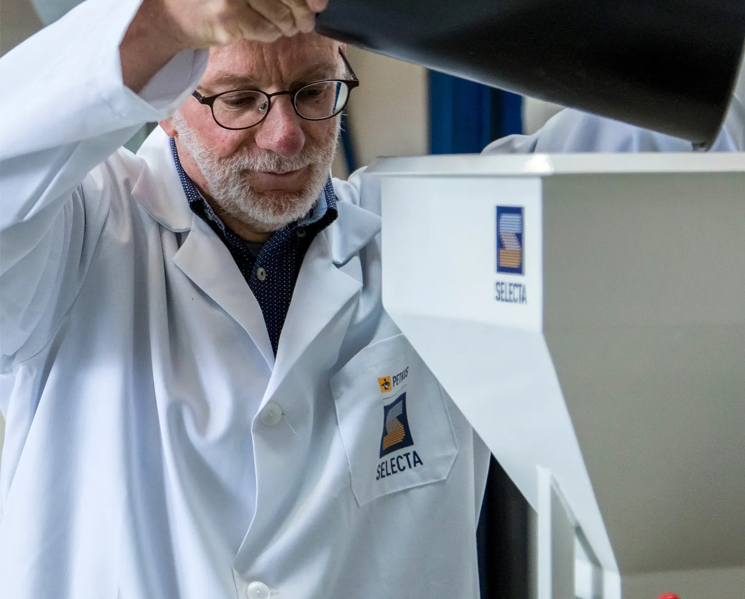 A man wearing glasses and a white lab coat with a 'SELECTA' logo operates a piece of lab equipment, lifting its black lid.