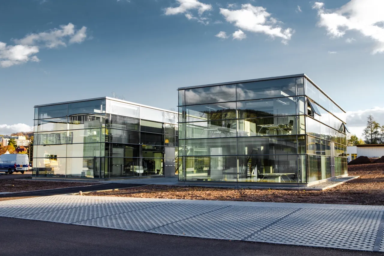 Two modern office buildings with glass exteriors reflecting the sky and surroundings, situated on a paved area under a partly cloudy blue sky.