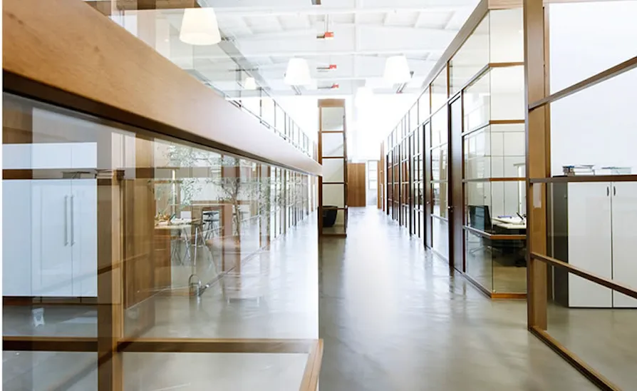 Bright modern office hallway with polished concrete floor and glass-walled offices framed with wood, extending into the distance under white ceiling lights.
