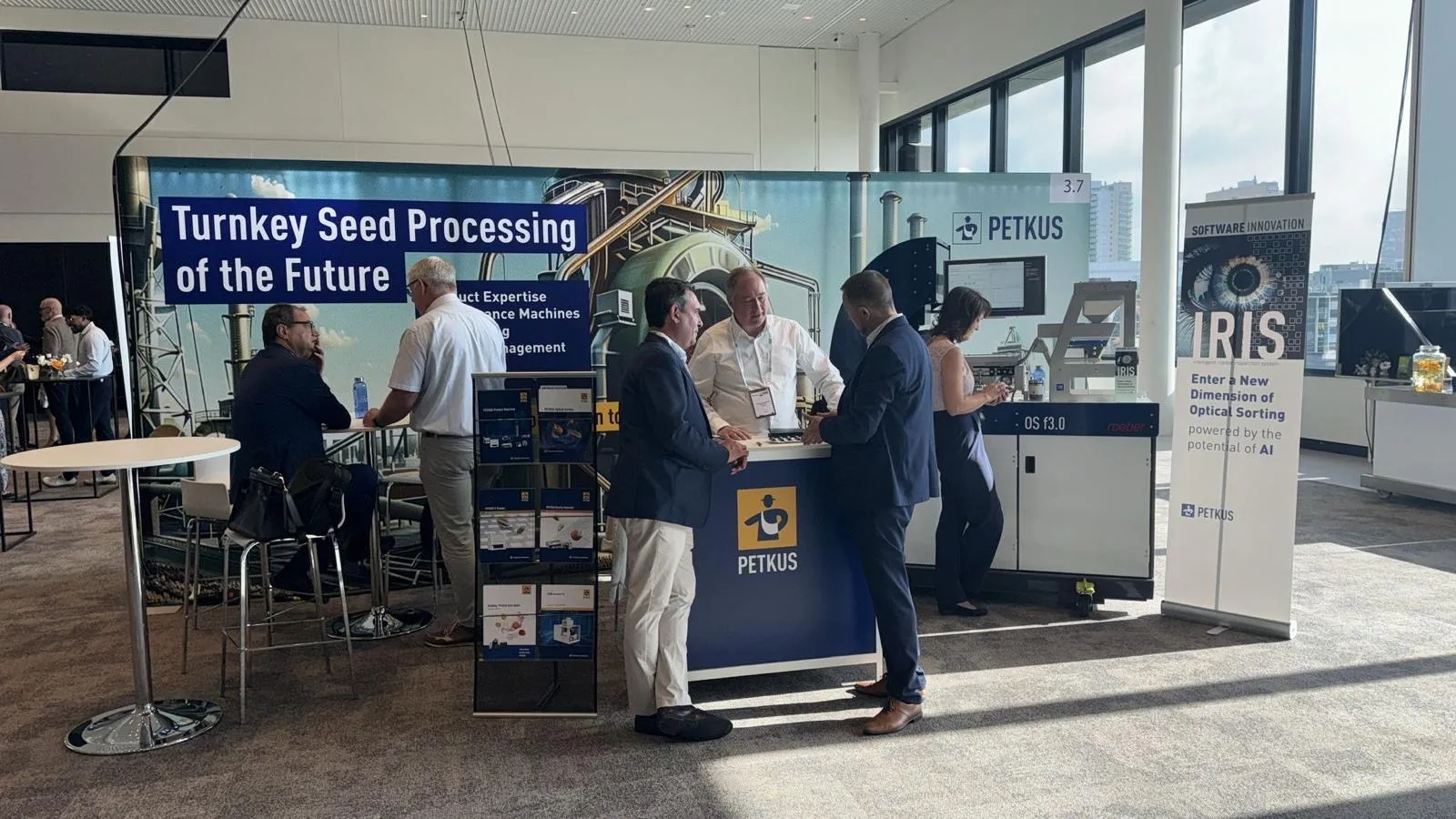 Trade show booth for PETKUS featuring a large banner reading 'Turnkey Seed Processing of the Future' with industrial equipment imagery. Four men in business attire are conversing at the booth counter, and a woman is engaged with a device near a machine labeled OS f3.0. A vertical banner promotes IRIS optical sorting software powered by AI. The setting is a bright indoor space with large windows and carpeted floor.