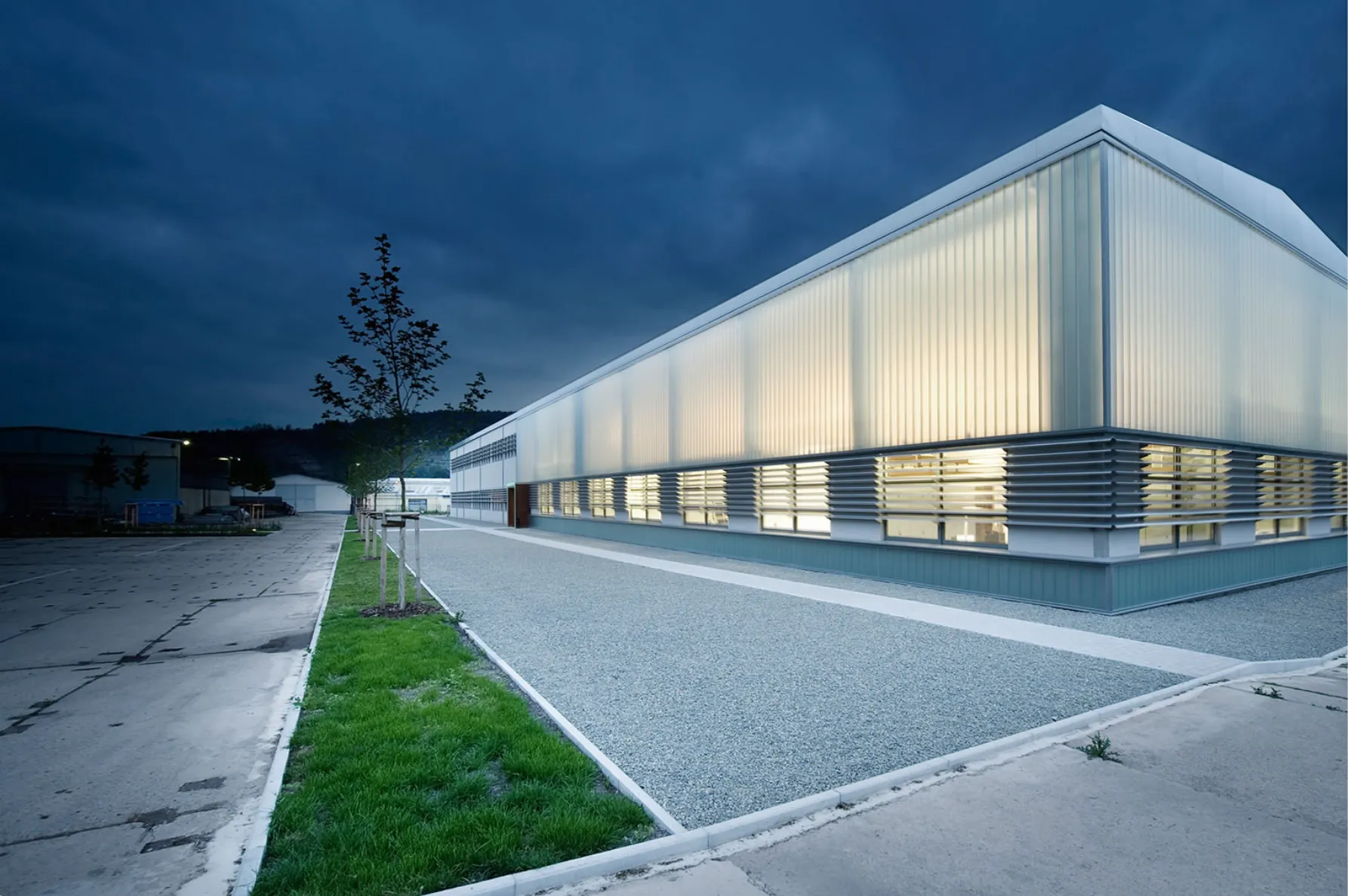 Modern industrial building at dusk with illuminated translucent upper facade, horizontal window slats on the lower level, gravel walkway, small trees planted along green strip, and adjacent paved area.