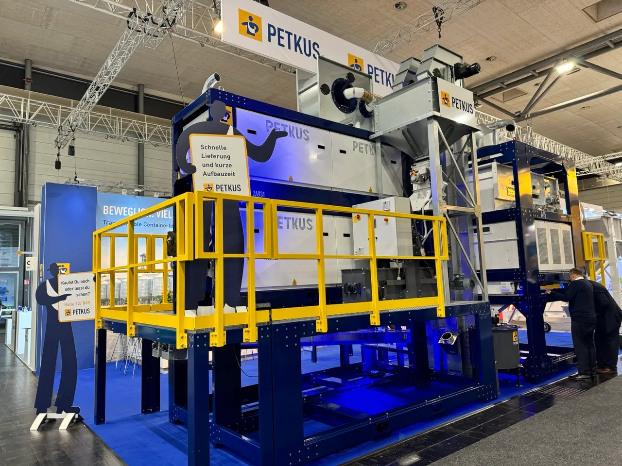 Large industrial machinery with yellow railings and PETKUS branding displayed in a trade show or exhibition hall, with two black silhouette figures holding signs in German and two people inspecting the equipment.
