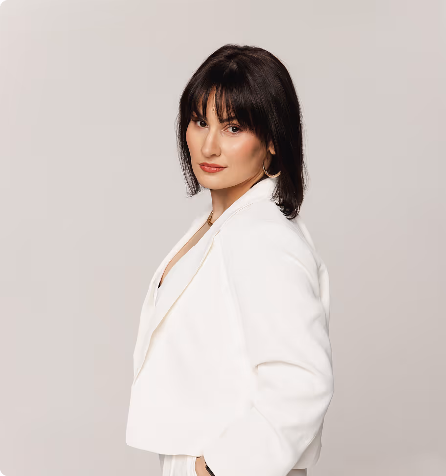 Portret kobiety w średnim wieku z ciemnymi włosami i delikatnym makijażem, ubranej w biały żakiet i białe spodnie, trzyma ręce w kieszeniach, stoi bokiem, twarz odwrócona w kierunku obiektywu, na jasnym tle