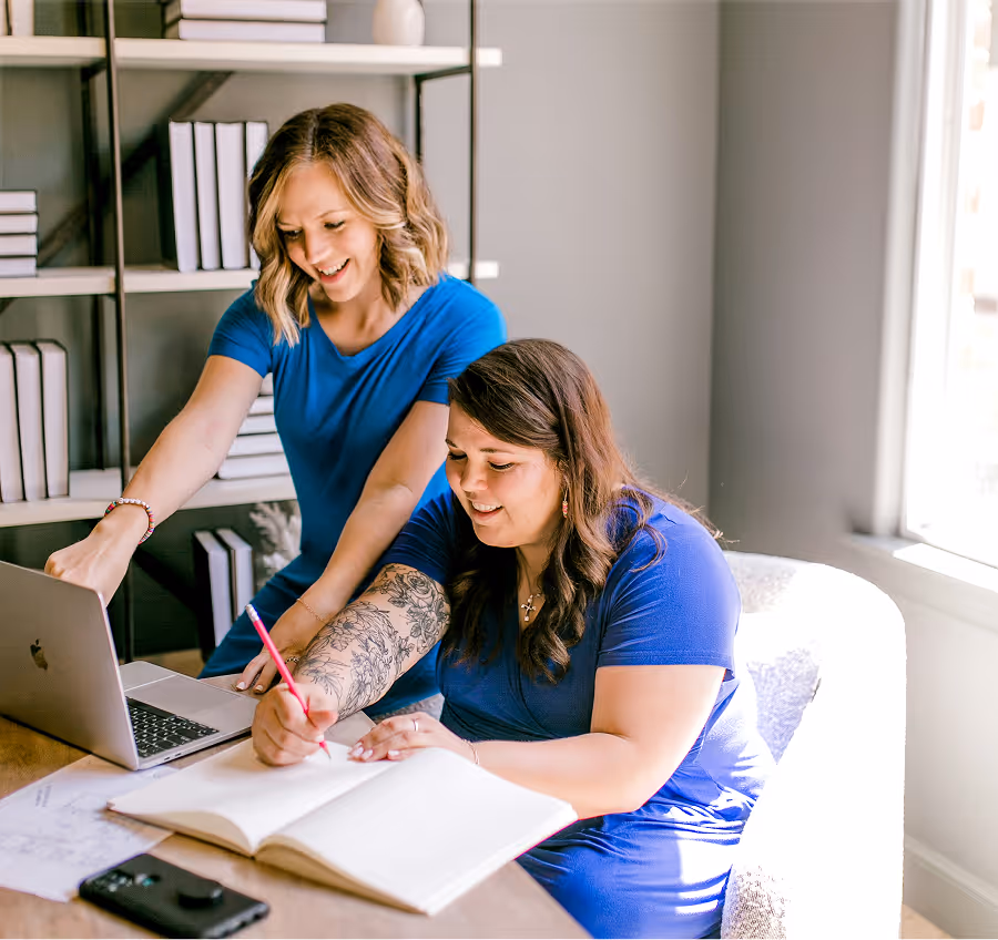 Dwie kobiety w niebieskich strojach, związane z fundacją lub stowarzyszeniem, pochylają się nad komputerem w jasnym wnętrzu