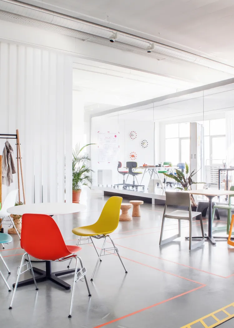 Bright modern office space with colorful chairs around white tables, indoor plants, and a glass-walled meeting room.